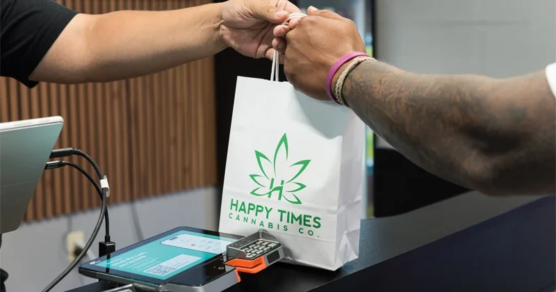 An employee handing a customer their purchase in a white bag with the Happy Times Cannabis logo on it.